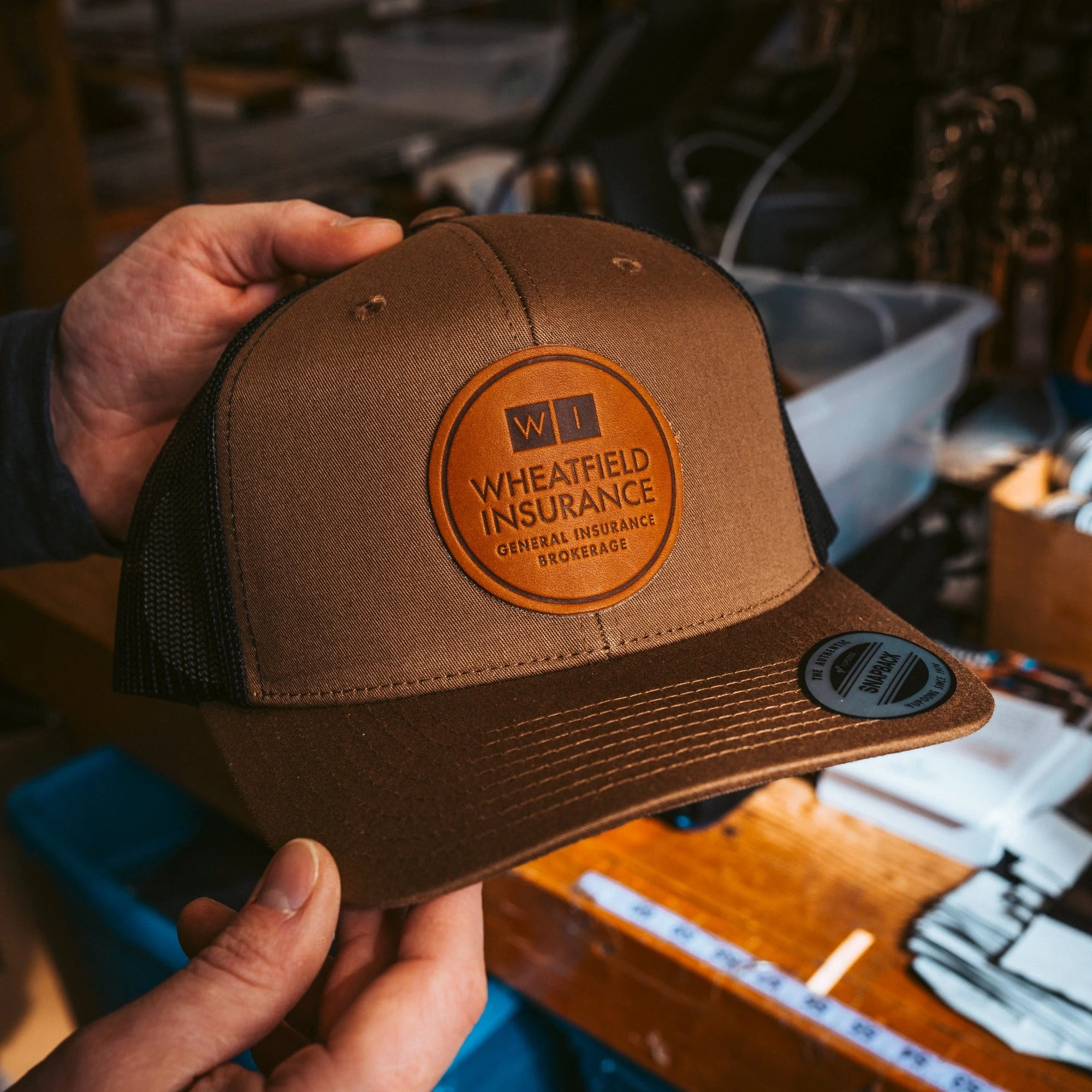 Hands holding a brown trucker hat with a stamped leather patch in the workshop