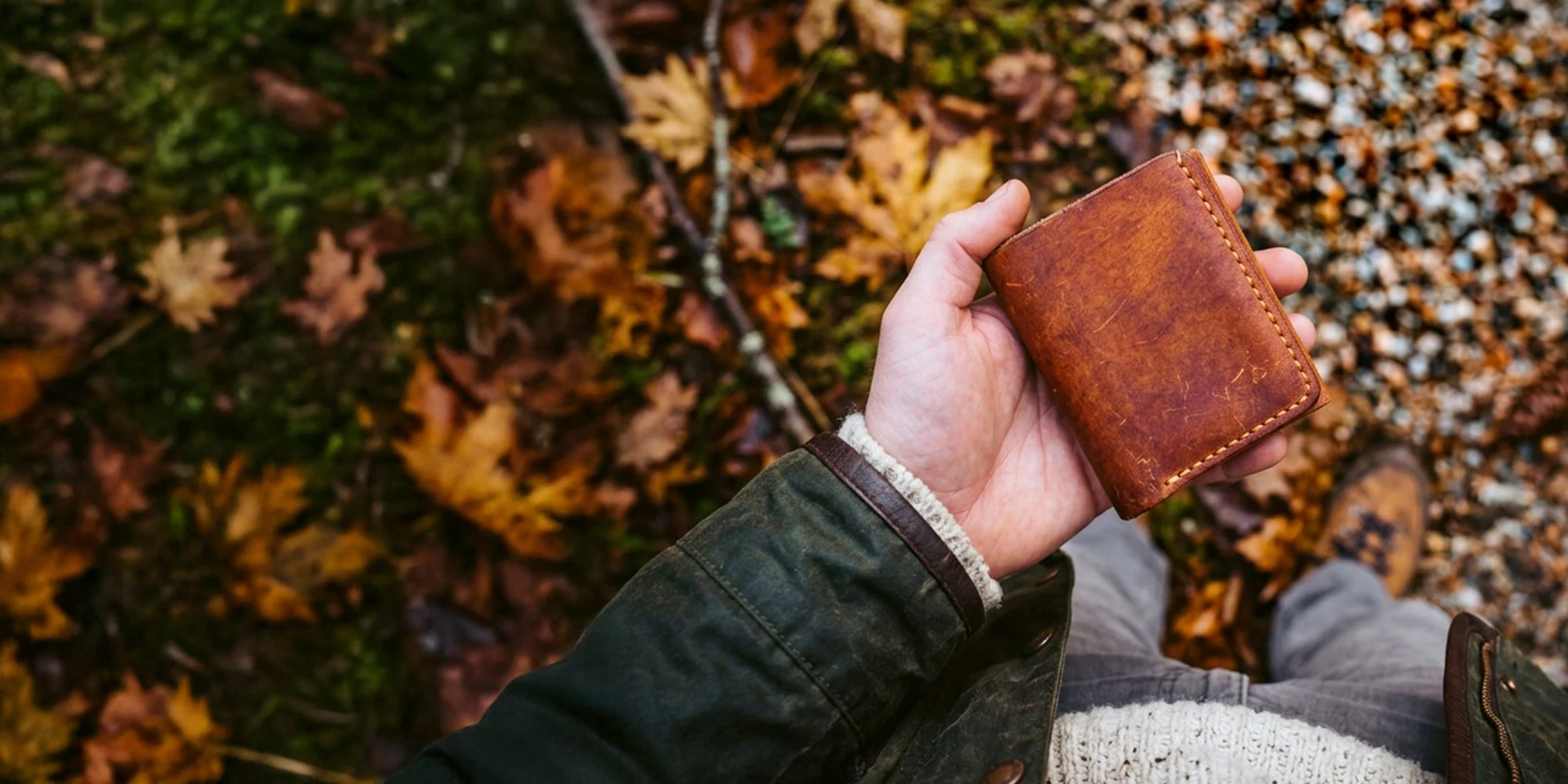 A hand holding a worn brown leather card wallet over autumn leaves