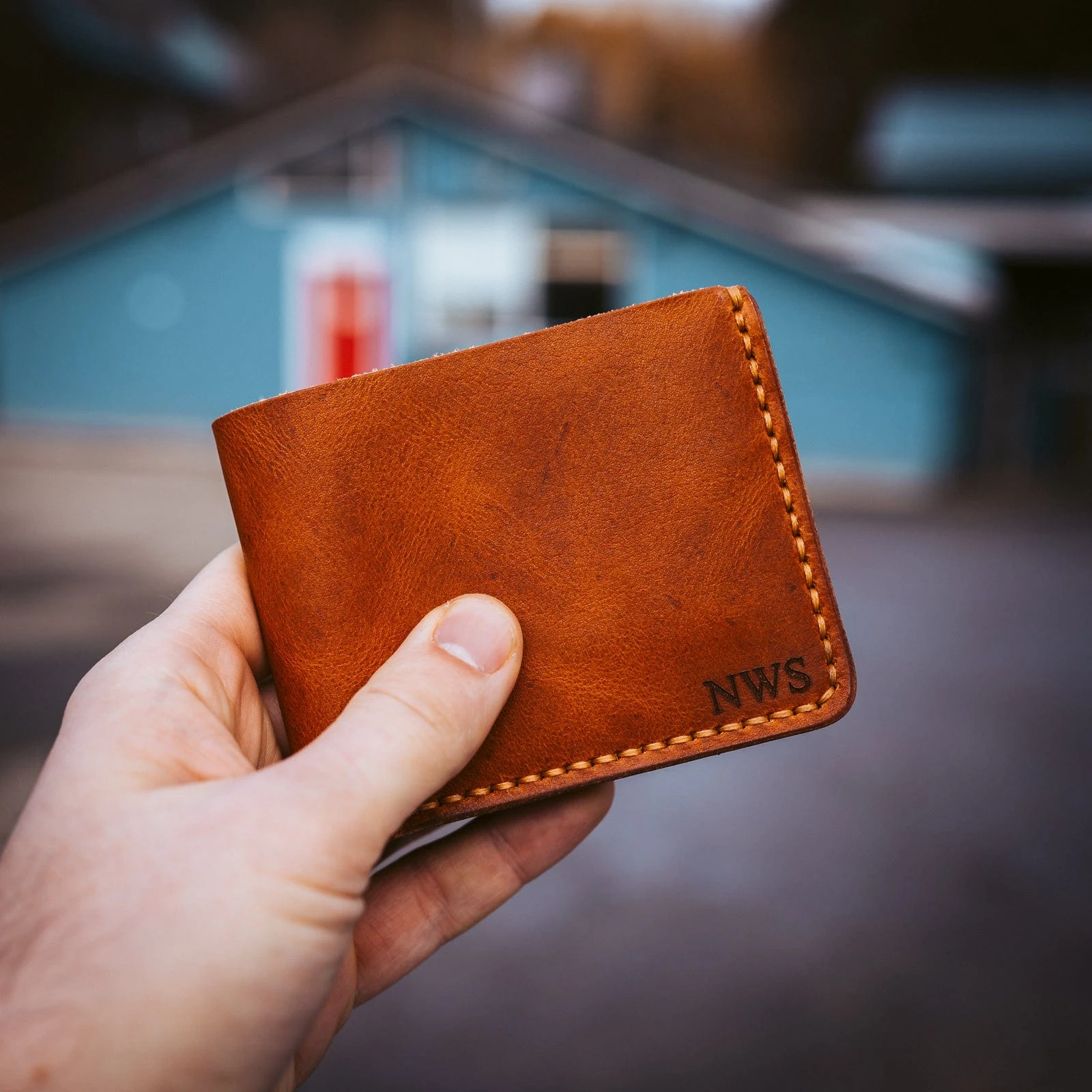 A hand holding a tan bifold wallet engraved with three initials in the bottom-right corner